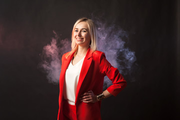 Fototapeta premium Young beautiful white blond girl in a bright red strict suit with a jacket and white blouse looks at the camera on a background of colored blue and pink smoke on a black isolated background