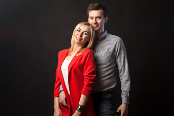 Obraz premium Young beautiful white blond girl in brightly red suit with jacket and white blouse and tall dark-haired guy hugging and kissing on isolated black background