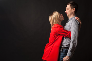 Young beautiful white blond girl in brightly red suit with jacket and white blouse and tall dark-haired guy hugging and kissing on isolated black background