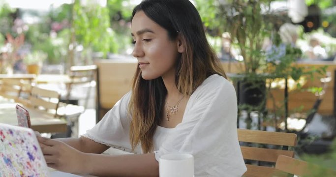 Young woman sitting at a shady table at an outdoor restaurant looking at her mobile phone with a pensive expression