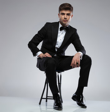 sexy confident man wearing a black tuxedo sitting