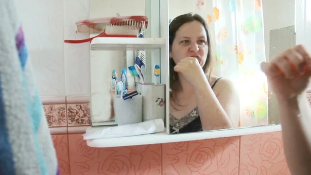 Overweight Long-haired Woman Brushing His Teeth In The Bathroom . Girl In Blue Nightgown Reflected In A Mirror Of The Locker. Long Hair Gathered In A Tail, With The Fallen Strands. Then She Wiped Out.