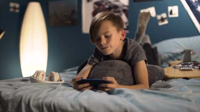 Teenager lying on bed with guitar and headphones and playing game on smartphone in leisure
