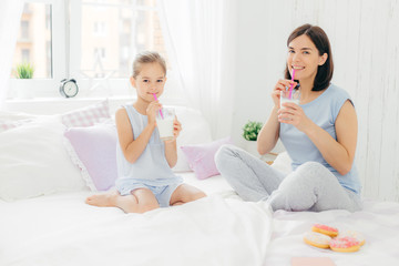 Obraz na płótnie Canvas Cheerful mother and daughter dressed in pyjamas, have breakfast in morning, drink milk shake with doughnuts, sit crossed legs on comfortable bed in white bedroom. People and lifestyle concept