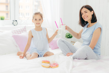 Obraz na płótnie Canvas Portrait of happy young mother and her small daughter hold glasses of milk shake, have breakfast in bed, look positively at camera, enjoys weekend at home and togetherness, dressed in nightwear