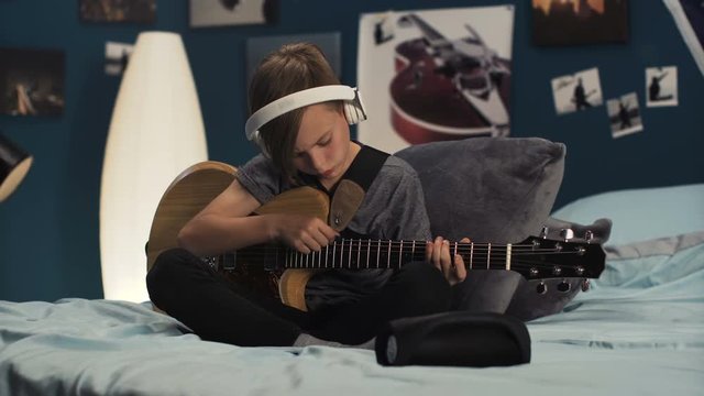 Teen boy sitting on bed at home and listening to music online in headphones while practicing guitar and watching video tutorial