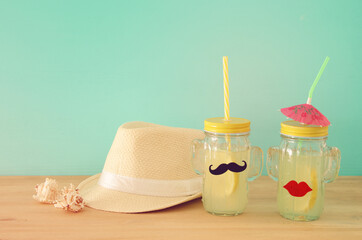 Image of fresh lemonade drink in cute cactus shape glasses wearing mustache and lips, over wooden table. Tropical summer romantic vacation concept.