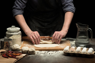 Chef hands are making a roll from dough on wooden board. black background