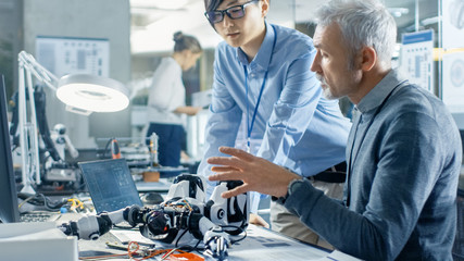 Two Engineers Talking while Working with Robot, Checking Data on Laptop Computer. In the Background Robotics Research Center Laboratory with Specialists Working.