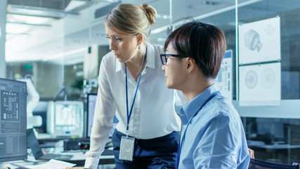 Asian Scientist Sitting at His Desk Consults Senior Engineer about Sophisticated Coding and Programming. In the Background Computer Science Research Laboratory with Robotic Arm Model.