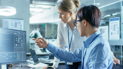Asian Scientist Sitting at His Desk Consults Senior Engineer about Sophisticated Coding and Programming. In the Background Computer Science Research Laboratory with Robotic Arm Model. © Gorodenkoff