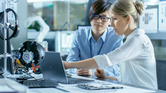 Asian Scientist Sitting At His Desk Consults Engineer About Sophisticated Coding And Programming. In The Background Computer Science Research Laboratory With Robotic Arm Model.