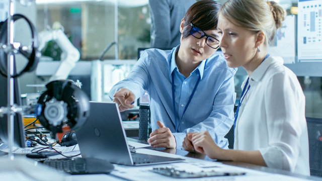 Asian Scientist Sitting at His Desk Consults Engineer about Sophisticated Coding and Programming. In the Background Computer Science Research Laboratory with Robotic Arm Model.