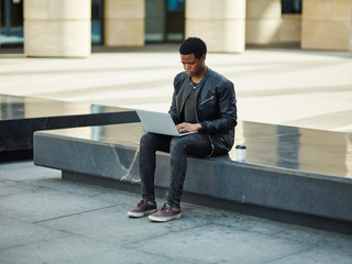 Young African man sitting on stone bench on city street and browsing Internet on laptop