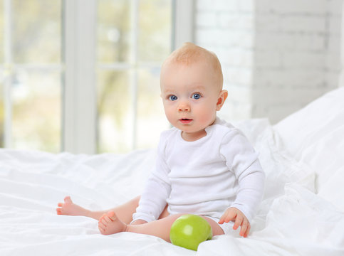 A beautiful baby sitting in a white bed with a green Apple.