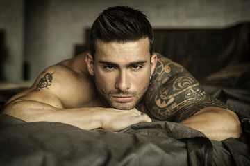 Shirtless muscular sexy male model lying alone on bed in his bedroom, looking at camera with a...