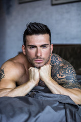 Shirtless muscular sexy male model lying alone on bed in his bedroom, looking at camera with a...