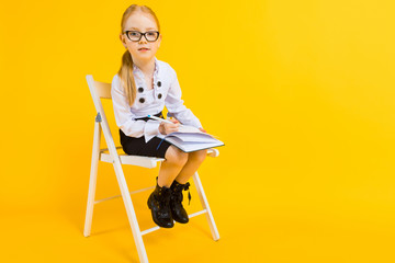 Fototapeta premium Girl with red hair on a yellow background. A charming girl in transparent glasses sits on a white chair and makes notes in a notebook.