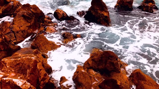Swirling white sea water breaking on a rocky coastal shoreline viewed high angle with brown rocks