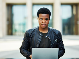 Portrait of confident African American businessman wearing leather jacket and looking at camera while working on laptop outdoors