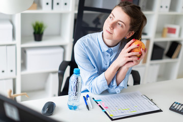 A young girl sits at a table in the office and holds an apple in her hand.