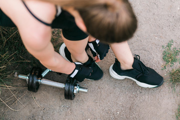 Close up of woman hands and legs standing on soil. She is leaning close to black dumbbells holding sneakers ties up the shoelaces