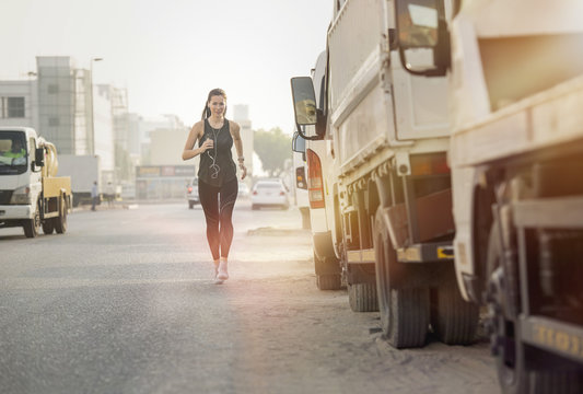 Athletic beautiful white female fitness model wearing long black sports wear with earphones connected to a mobile phone running or jogging in a urban street with trucks and sun flare in the background
