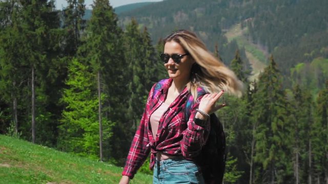 A young caucasian woman with a backpack and sunglasses rises up the mountain, adjusts the hair that develops in the wind. The concept of personal growth, overcoming difficulties in business