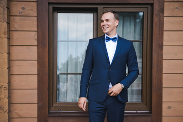 Morning of the groom. The groom smiles in a blue suit and a bow tie. Outdoor. A wooden brown house.