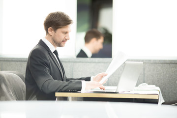 Young analyst or financier with papers working in front of laptop by table