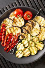 Grilled vegetables: eggplant, zucchini, pepper. Skewer with mushrooms. On a black plate and a black background. Toned.