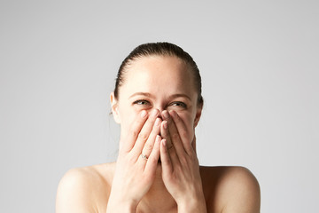 Beautiful brunette girl with naked shoulders laughs with closes her face two hands.Model with light nude make-up, gray studio background.Medical and cosmetic facial skin healthcare concept