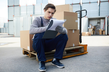 Young man in workwear sitting on cart with load and filling working documents outdoors