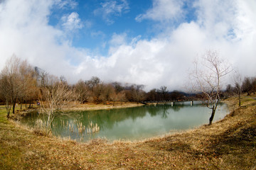 Forest lake with bridge during the sunny day with winter trees and blue cloudy sky. Beautiful natural mountain lake with forest in the background and stormy clouds on the sky.