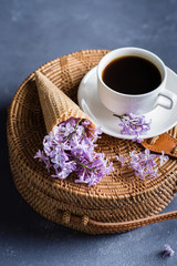 Fashionable rattan bag, cup of coffee and purple lilac in waffle cone on blue concrete background. Spring Summer Card Concept. Top view