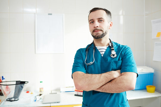 Young doctor in uniform crossing his arms on chest while looking at you in his office