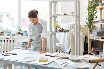 Creative young woman in workwear leaning over table and sketching in her studio