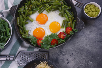 Fried eggs in an old pan with tomatoes, avocado pesto, green beans, pepper and salad on a dark background