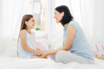 Lovely mother and daughter enjoy togetherness, keep hand together, have pleasant talk, sit on comfortable bed in morning, sit in light bedroom, have good relationships. Family and love concept
