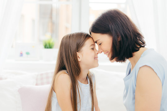 Close up shot of affectionate young mother and her little daughter touches noses and enjoy togetherness, pose against white background and bedroom interior, smile gladfully. Motherhood concept