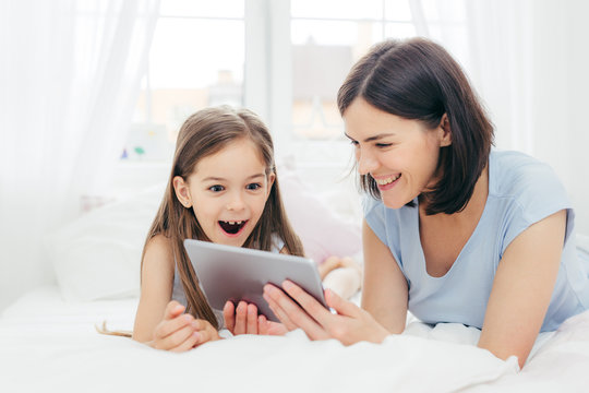 Beautiful mother and daughter watch something funny on tablet computer, connected to wireless internet, spend free time in bedroom, have surprised happy expression. People and leisure concept
