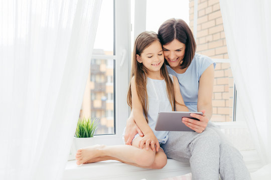 Horizontal shot of beautiful brunette mother and daughter spend free time together, sit on window sill, watch interesting movie via digital tablet, being in bedroom. People, technology, family concept