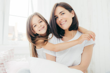 Portrait of happy Caucasian mother with charming smile and her small daughter embraces with love...