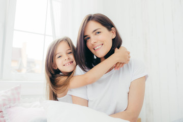 Indoor shot of good looking brunette mother with gentle smile and her small daughter gives hug, enjoy domestic atmosphere, pose against spacious white bedroom interior. Motherhood and family concept