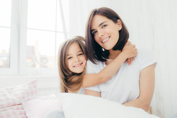 Obraz na płótnie Canvas Positive daughter and mum embrace each other, being in high spirit after good sleep, pose in bedroom, smile gently on face, have pleasant appearance. Pretty child with her mother. Morning and bed time