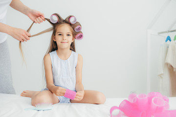 Obraz na płótnie Canvas Attractive female child has curlers on head, going to have wonderful hairstyle, recieves beauty lessons from affectionate mother, isolated over white background. Children and beauty concept.