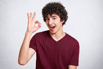 Everything will be fine! Satisfied curly man looks with assured and happy expression, shows ok sign, demonstrates his readiness and awareness, dressed in casual t shirt, isolated over white background
