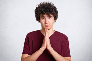 Please, forgive me! Handsome young curly male makes praying gesture, keeps palms pressed together,...