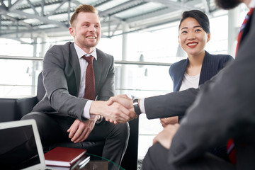 Smiling bearded entrepreneur wearing elegant suit sitting opposite his business partner and shaking his hand after successful completion of negotiations, pretty Asian assistant manager