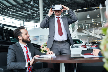 Smiling client wearing classical suit using VR headset while testing new car, two dealers sitting...